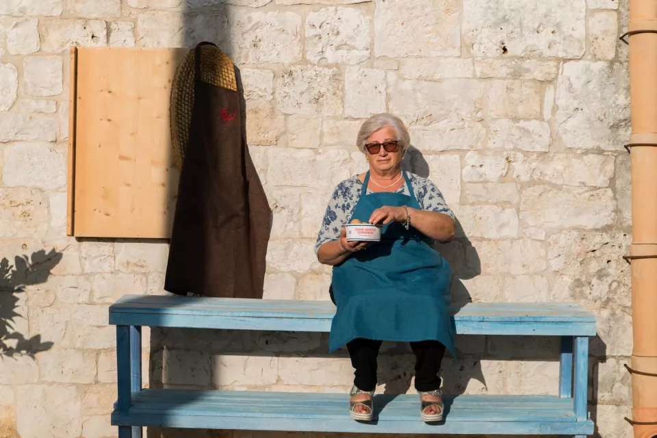 Una nonna seduta su una panca azzurra davanti a un muro di pietra, con una ciotola in mano
